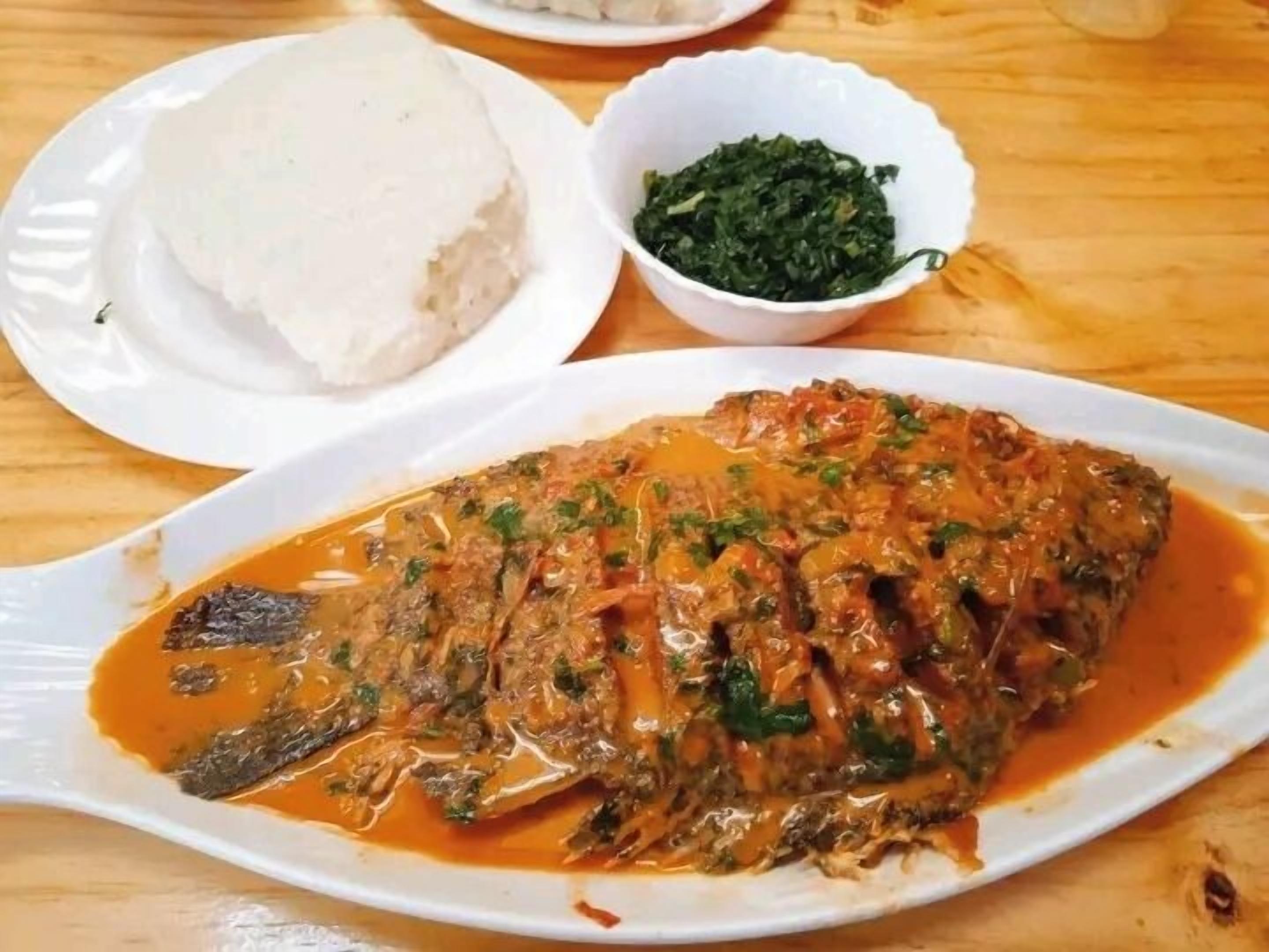 A plate of fish in sauce, a bowl of greens, and a plate of white food on a wooden table.