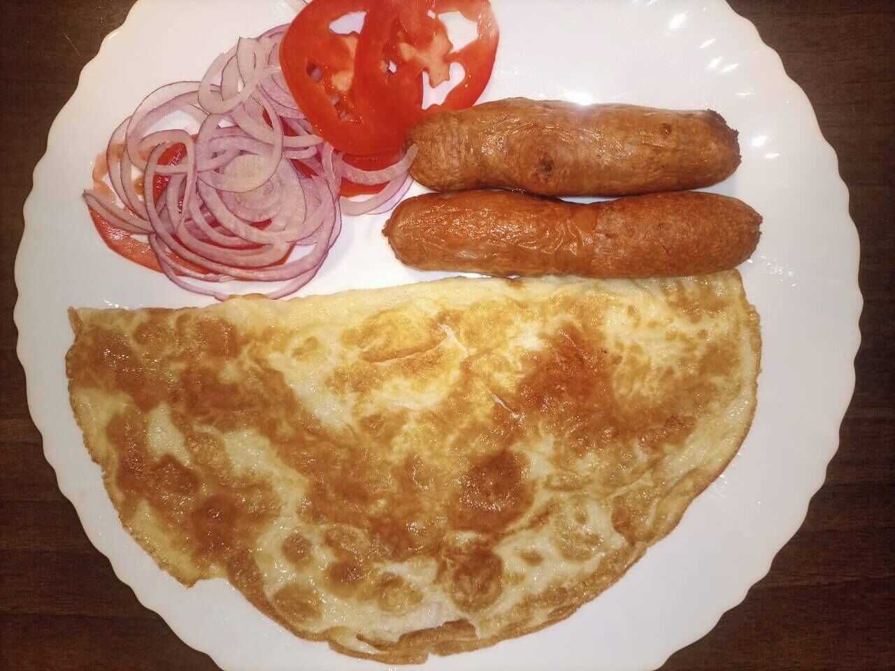 A plate with an omelette, two sausages, and sliced onions and tomatoes.