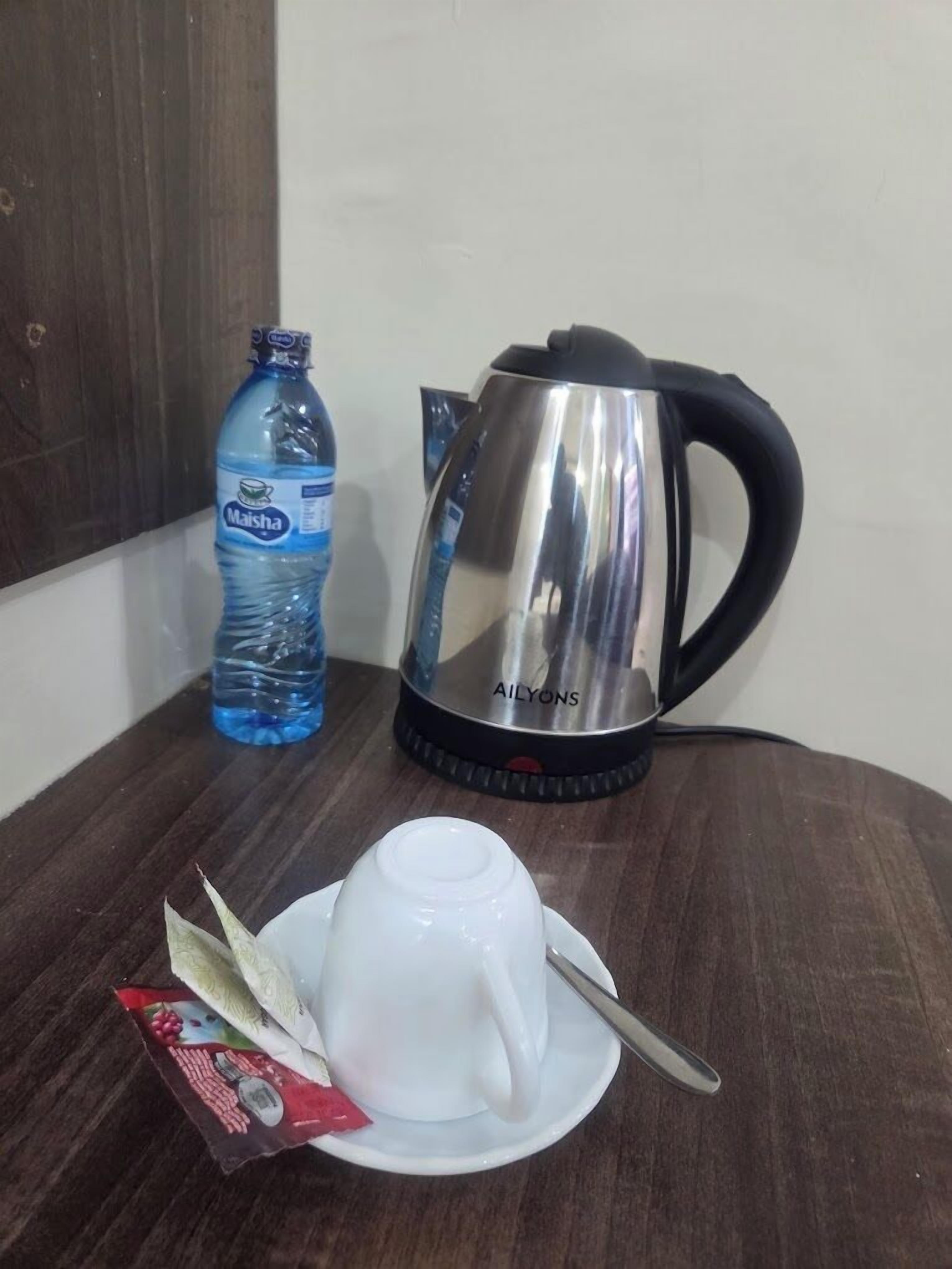 A white cup with a spoon on a saucer, a stainless steel kettle, and a water bottle on a wooden table.