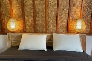 Una cama con dos almohadas blancas y una colcha oscura, frente a un cabecero de bambú trenzado con dos lámparas de pared.