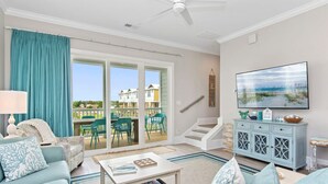 Un salon spacieux doté d’une grande porte-fenêtre coulissante donnant sur un balcon, d’un ventilateur de plafond, d’un téléviseur à écran plat fixé au mur et d’une décoration aux tons bleu clair.
