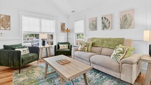 A spacious living room with a green sofa, a beige sofa, a wooden coffee table, and a green armchair. There are large windows, a lamp, and wall art.