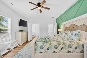 Un dormitorio con cama, ventilador de techo, televisor, cómoda y ventana con persianas.