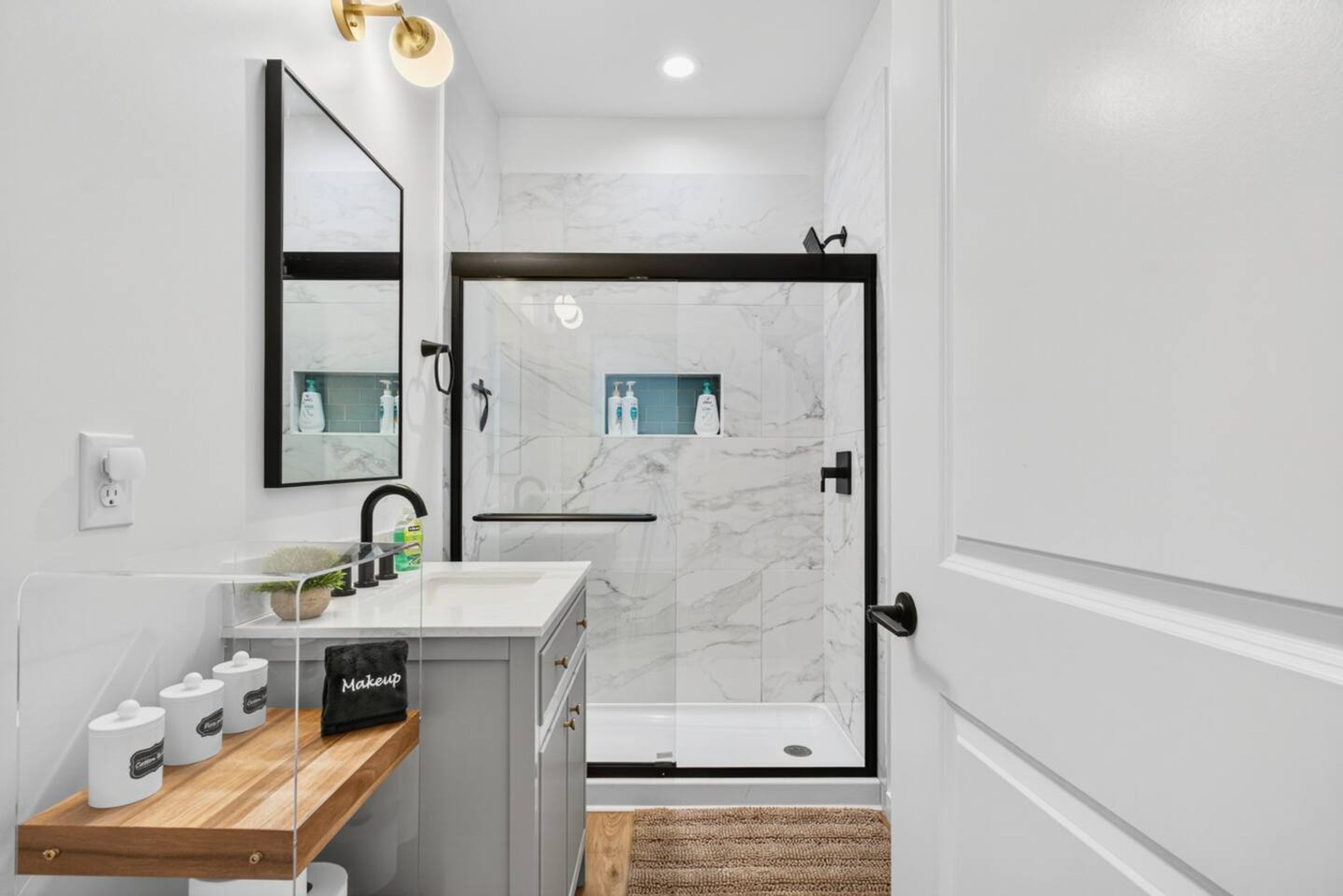 A modern bathroom with a glass shower enclosure, a white sink with a mirror above, and a wooden shelf with storage containers.
