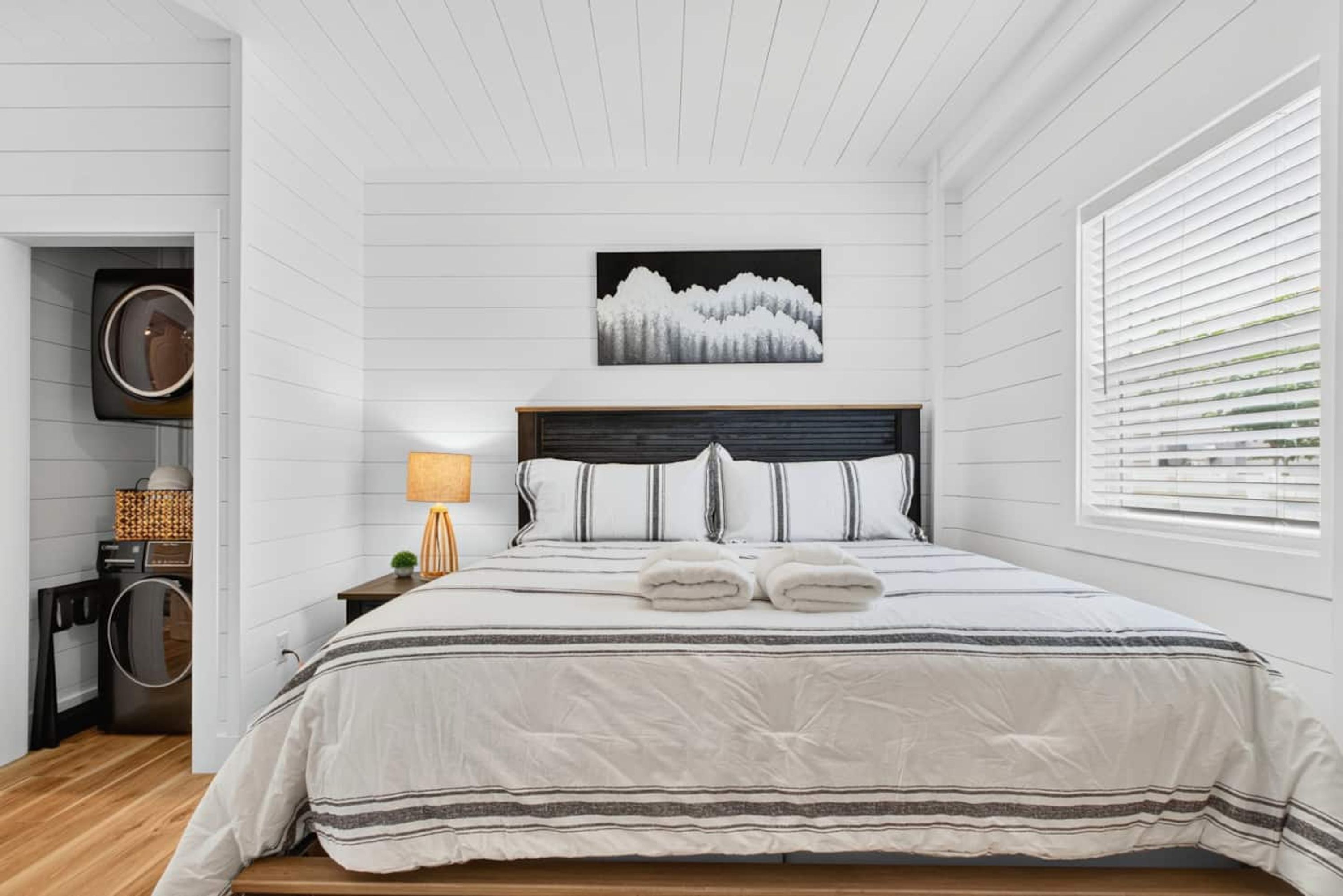 Quarto com cama, mesa de cabeceira, luminária, quadro e janela com persianas.