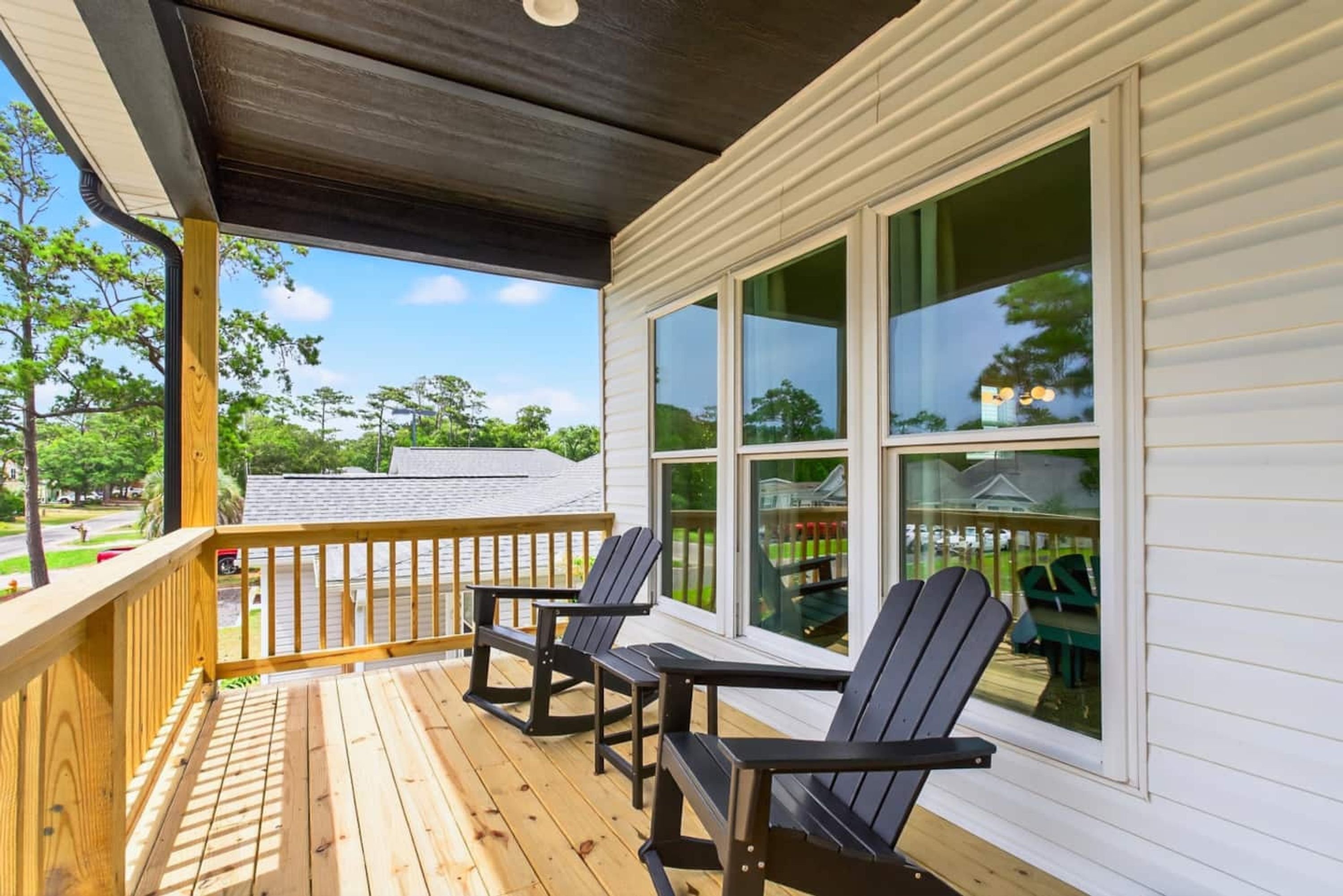 Una terraza de madera con dos sillas Adirondack y una mesa, con vista a un patio trasero con sillas verdes y una valla.