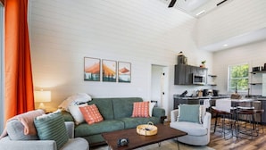 Uma sala de estar moderna com um sofá verde, poltronas cinzas e uma mesa de centro de madeira. Uma cozinha com micro-ondas e armários é visível ao fundo.