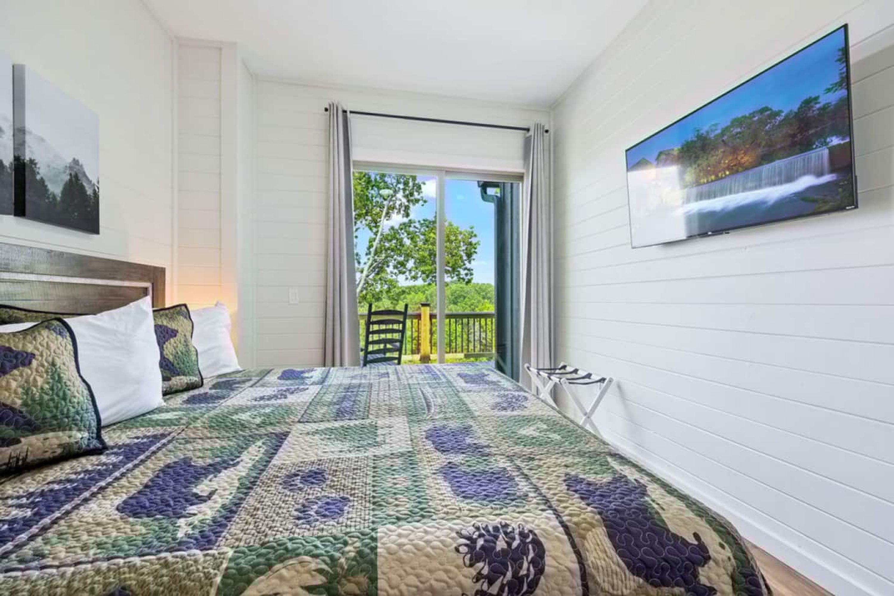 A bedroom with a large bed, a TV mounted on the wall, and a sliding glass door leading to an outdoor area.