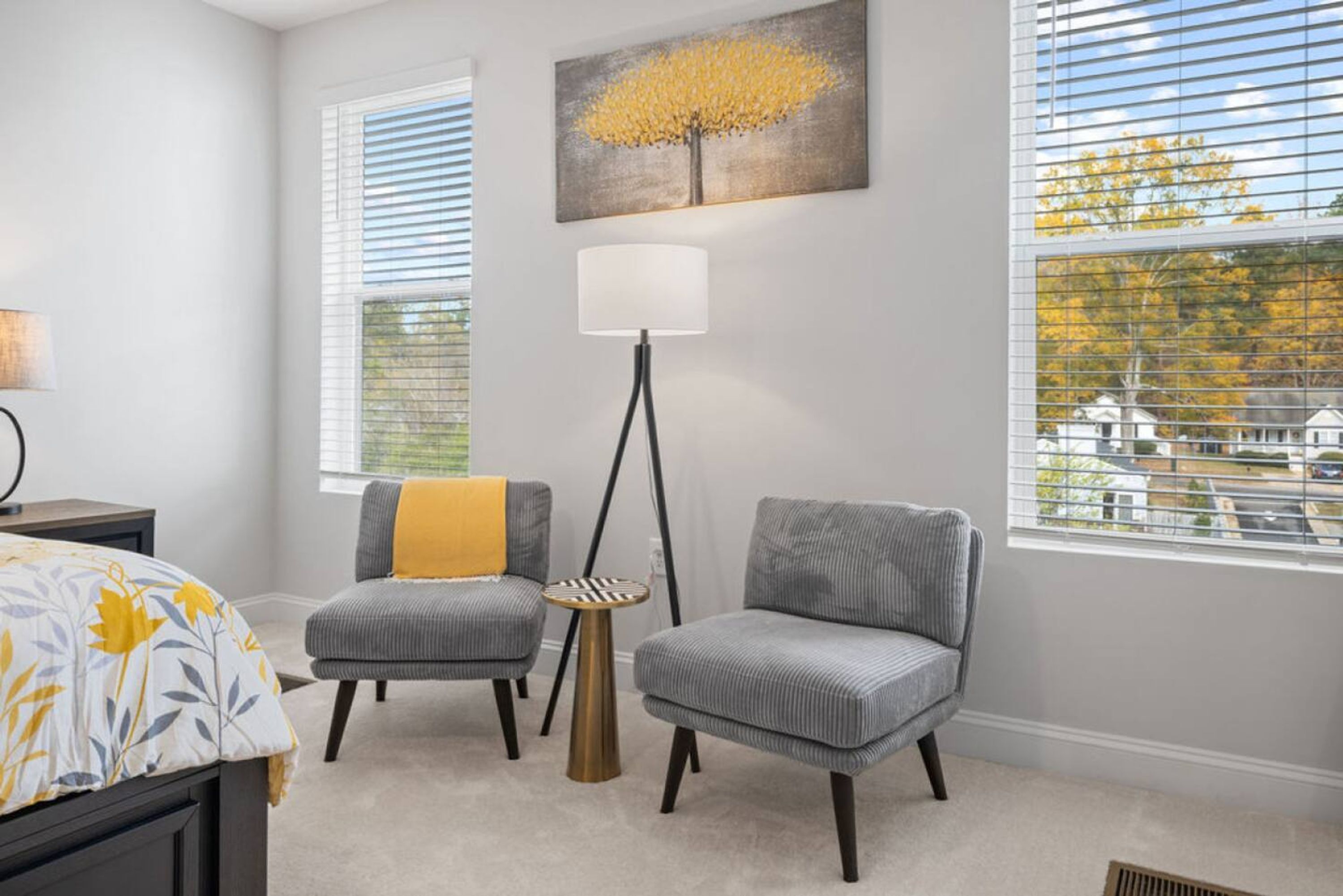A bedroom with two gray armchairs, a floor lamp, a bed with a yellow and white floral bedspread, and a painting of a tree on the wall.