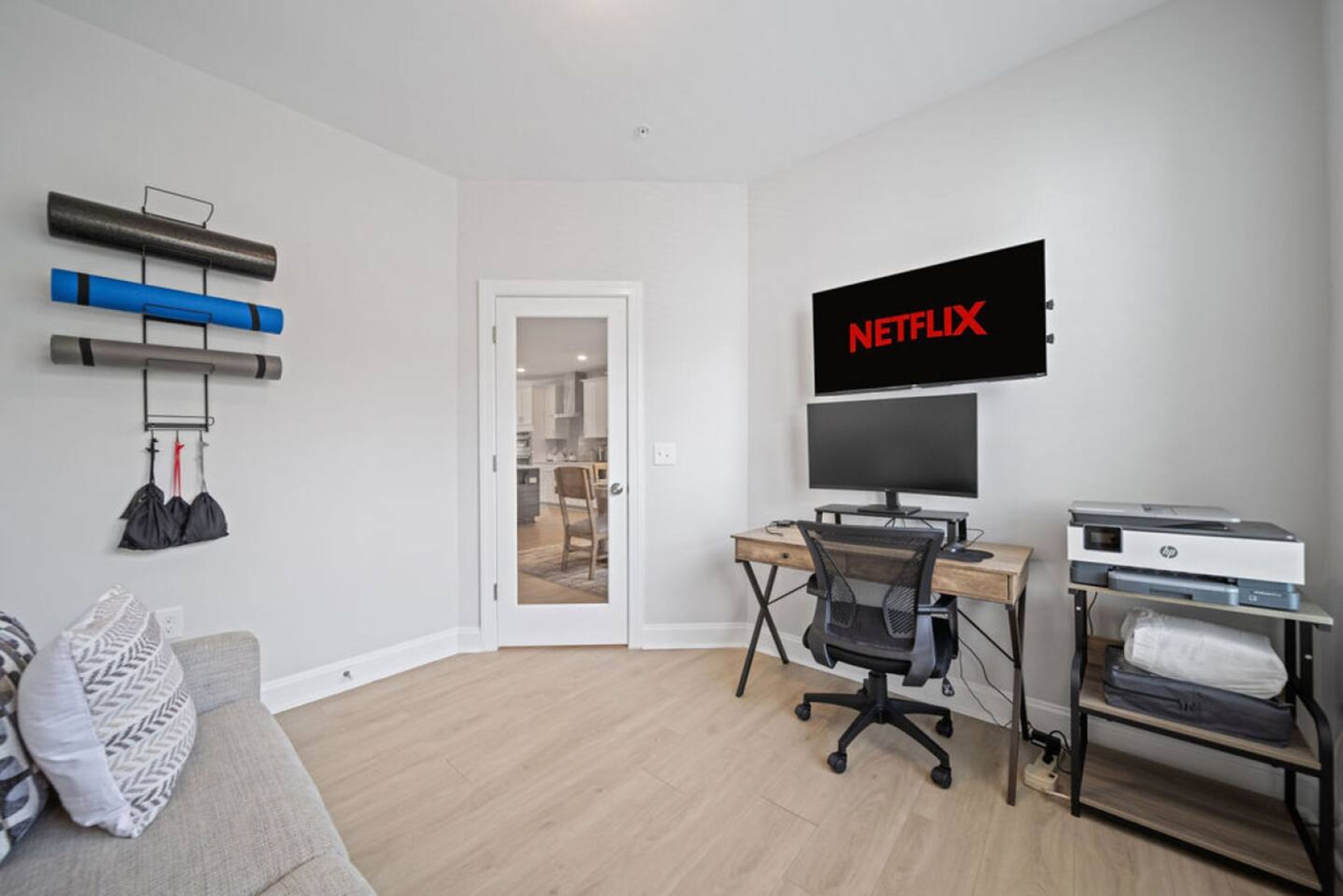 A modern living room with a grey sofa, a wooden desk with a computer, a printer, and a mounted TV displaying the Netflix logo.