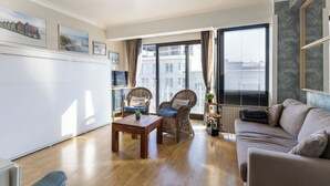 A living room with a wooden floor, a sofa, two wicker chairs, a coffee table, and a large window with blinds. - 1 bedroom accommodation in Sint Idesbald (Sint Idesbald)
