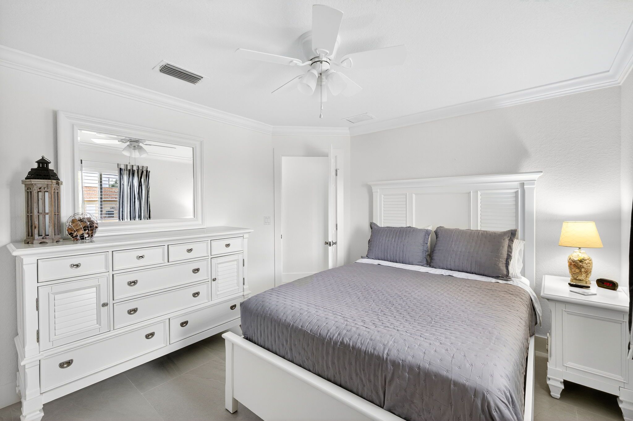 Une chambre à coucher avec un lit blanc, une table de chevet avec une lampe, une commode, un ventilateur de plafond et une fenêtre avec des rideaux.