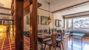 Un área de comedor con una mesa y sillas de madera, un sofá, y vistas a un pueblo montañoso a través de ventanas grandes.