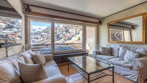Un salón moderno con una mesa de centro de vidrio, dos sofás y grandes ventanales con vistas a un pueblo montañoso.