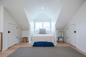 A bedroom with a sloped ceiling, a bed with a white and blue bedspread, a chair, and a woven basket on the floor. - The Kinsey, 3BR @ Plein Air by Velvet Ditch Villas (Taylor)