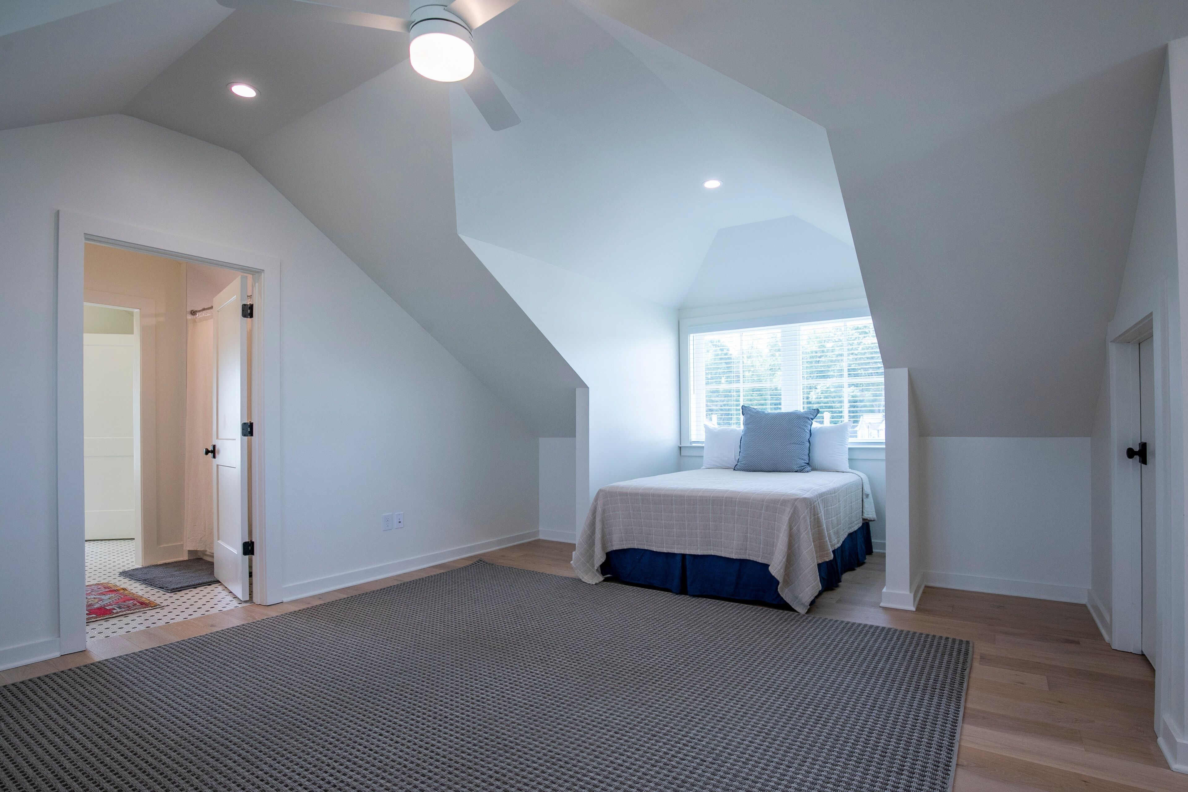 A bedroom with a sloped ceiling, a bed with blue bedding, a window with blinds, and a door leading to another room.