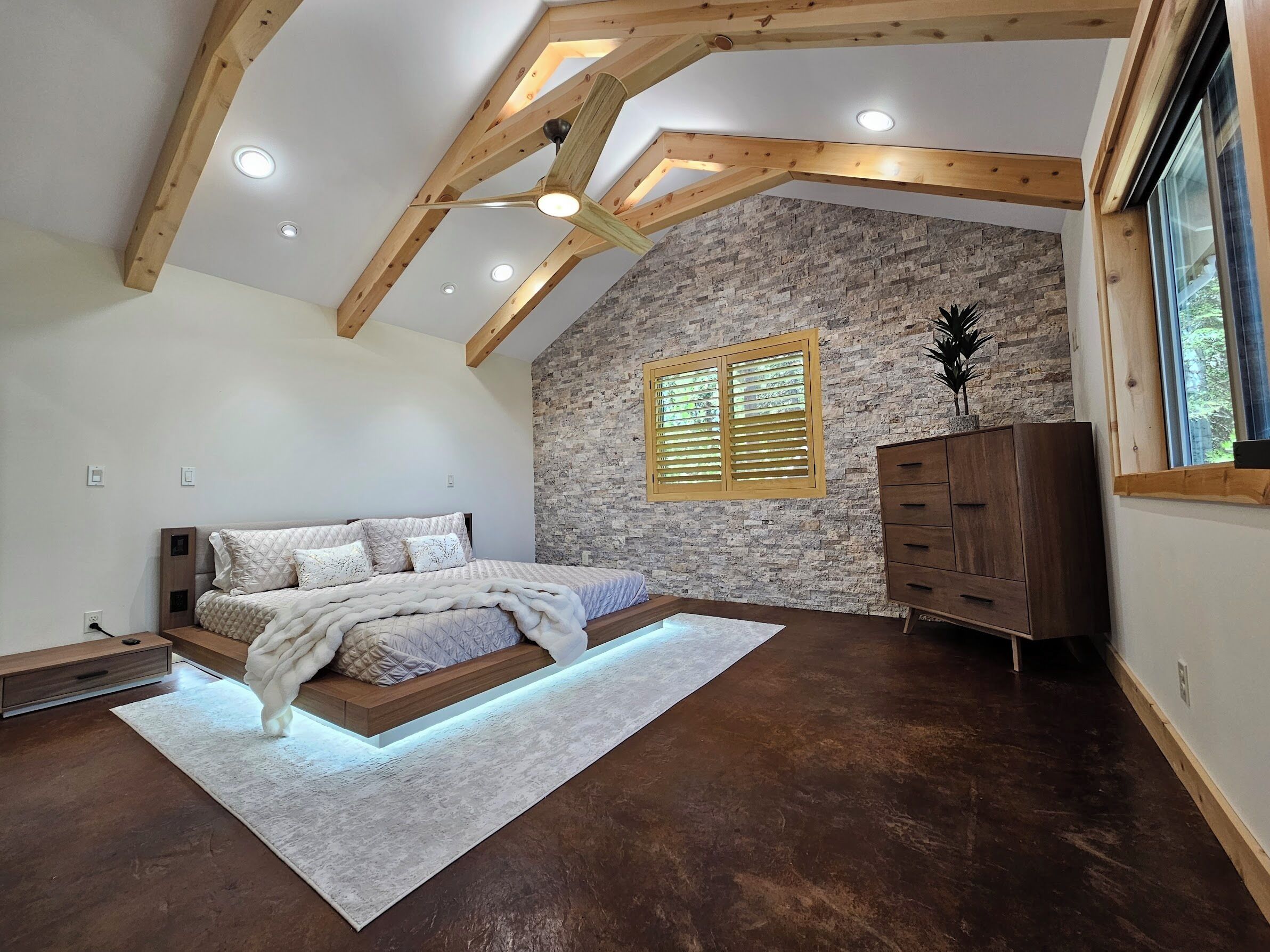 Une chambre avec un plafond en pente, des poutres apparentes, un mur d’accent en pierre, un lit recouvert d’une couvre-lit à motifs blancs et gris, une commode en bois et une fenêtre avec des stores.