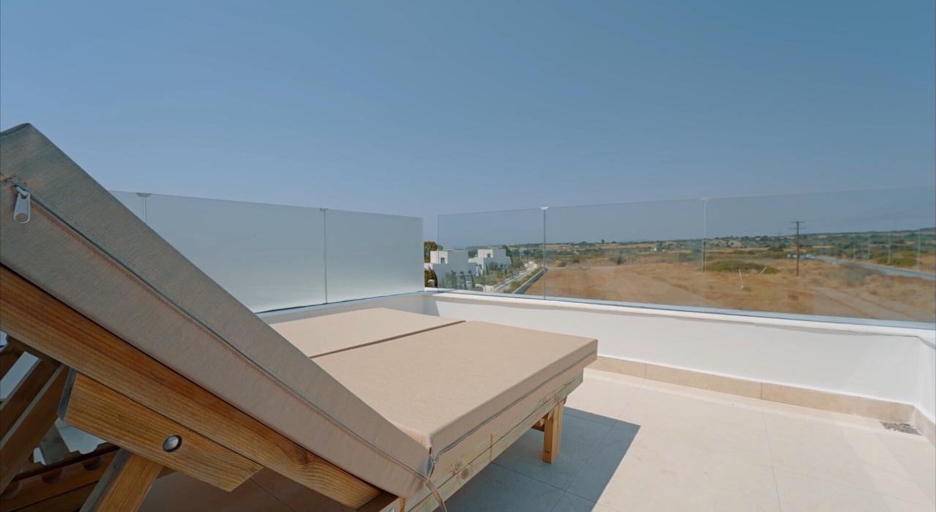 Un balcón con una silla de madera y un colchón, con vistas a un paisaje llano y edificios a la distancia.