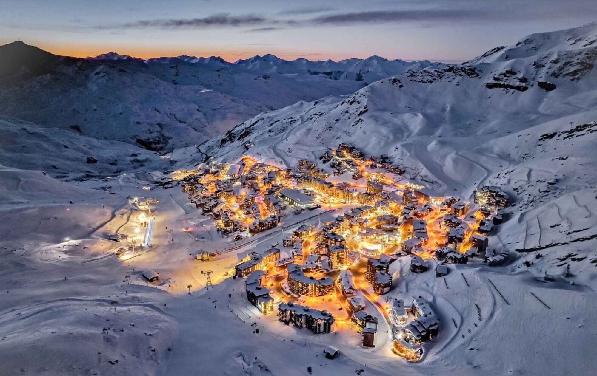 A snow-covered mountain town at dusk with illuminated buildings and winding roads.