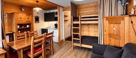 Une pièce compacte et moderne avec un plancher en bois, un coin repas avec une table et des chaises en bois, un lit superposé avec une échelle, un canapé et un coin cuisine équipé d’un évier et de placards.