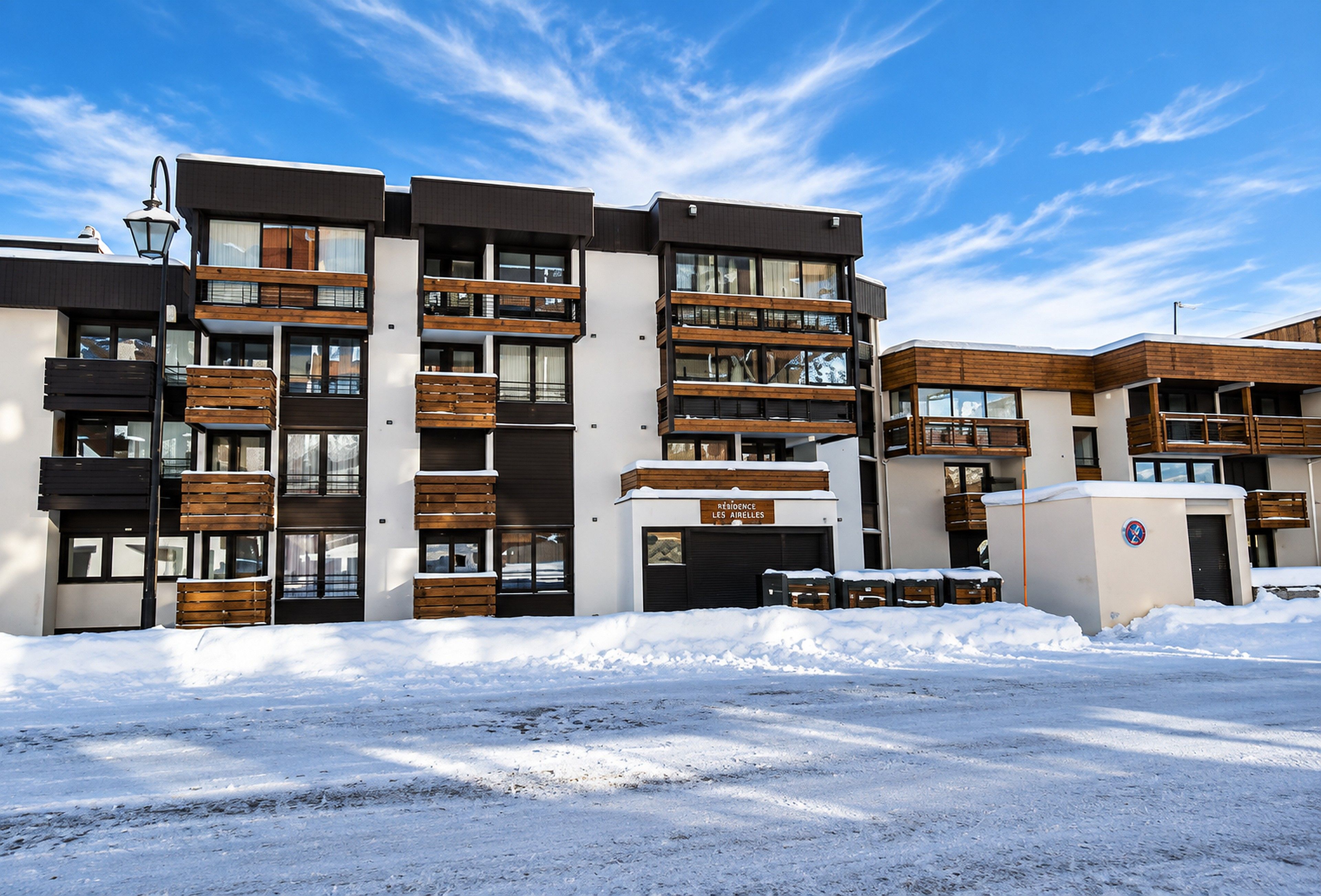 A modern multi-story building with balconies, a sign indicating 'Residence Les Airelles,' and a snow-covered ground.