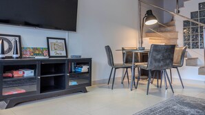A modern living room with a flat-screen TV, a bookshelf, a dining area with chairs, and a staircase.
