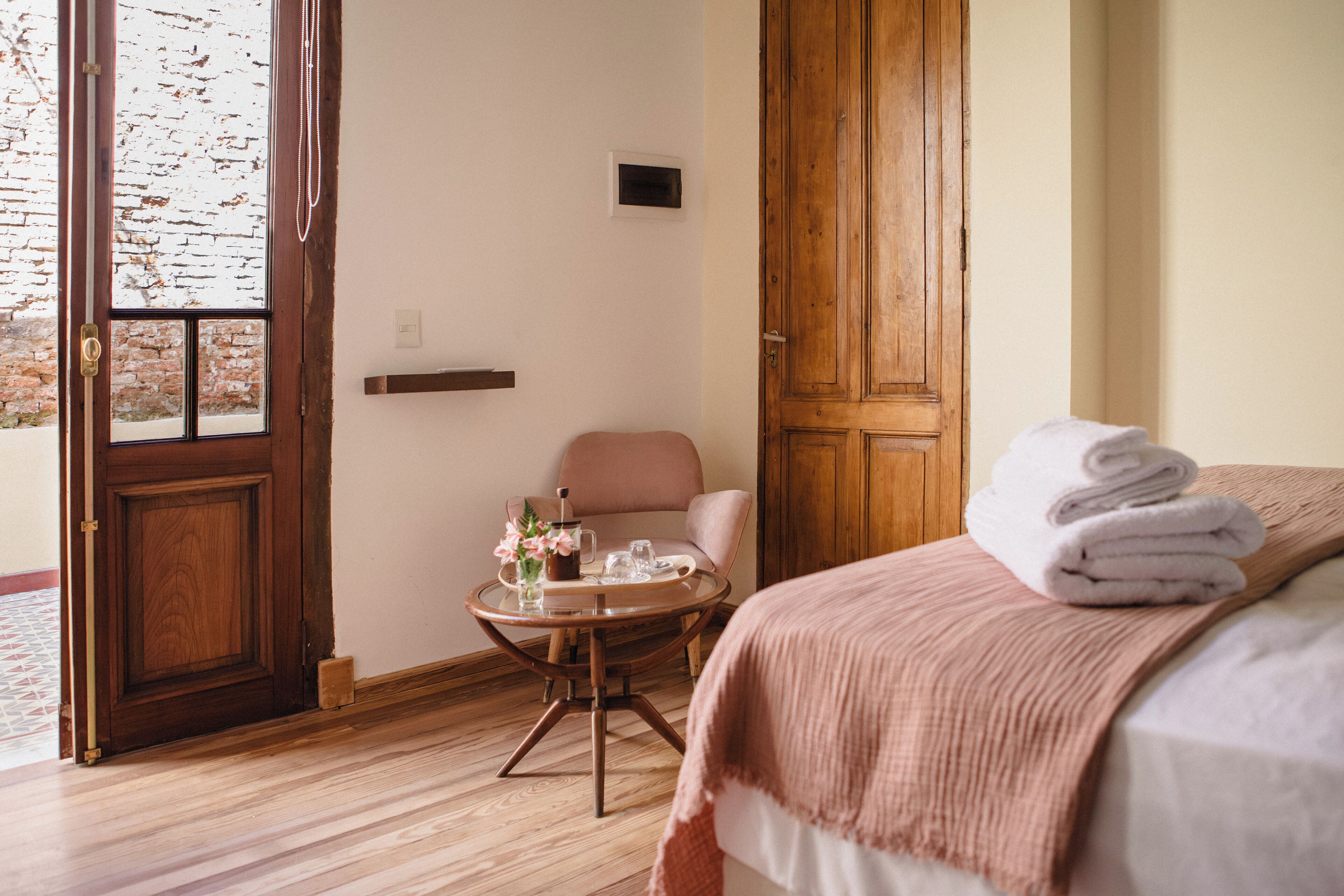 A hotel room with a wooden door, a round table with a vase and glasses, a chair, and a bed with a rolled towel.