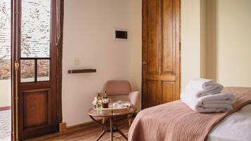 A hotel room with a wooden door, a round table with a vase and glasses, a chair, and a bed with a rolled towel.