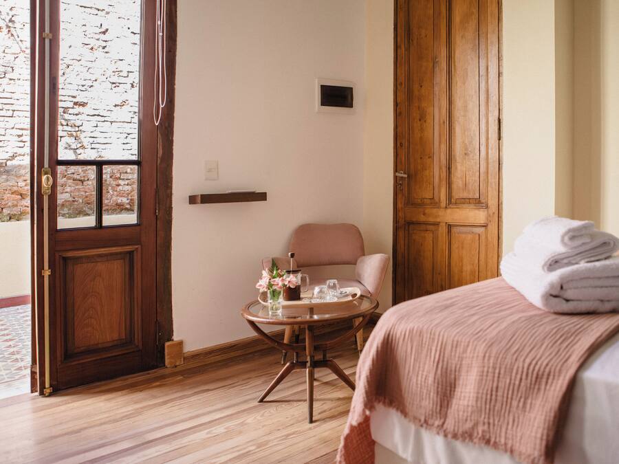A hotel room with a wooden door, a round table with a vase and glasses, a chair, and a bed with a rolled towel.