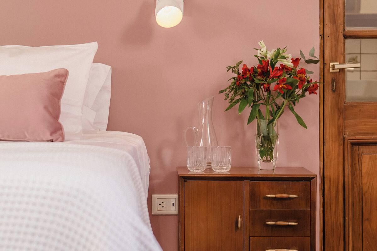 Een slaapkamer met een bed, nachtkastje, vaas met bloemen en een houten deur.