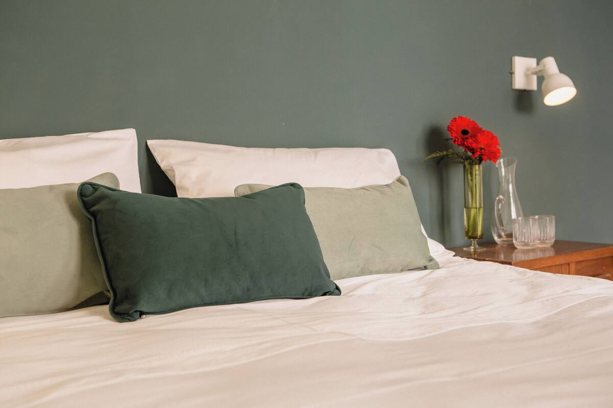 Een netjes opgemaakt bed met witte en groene kussens, een vaas met rode bloemen, en een nachtkastje met een glas en een lamp.
