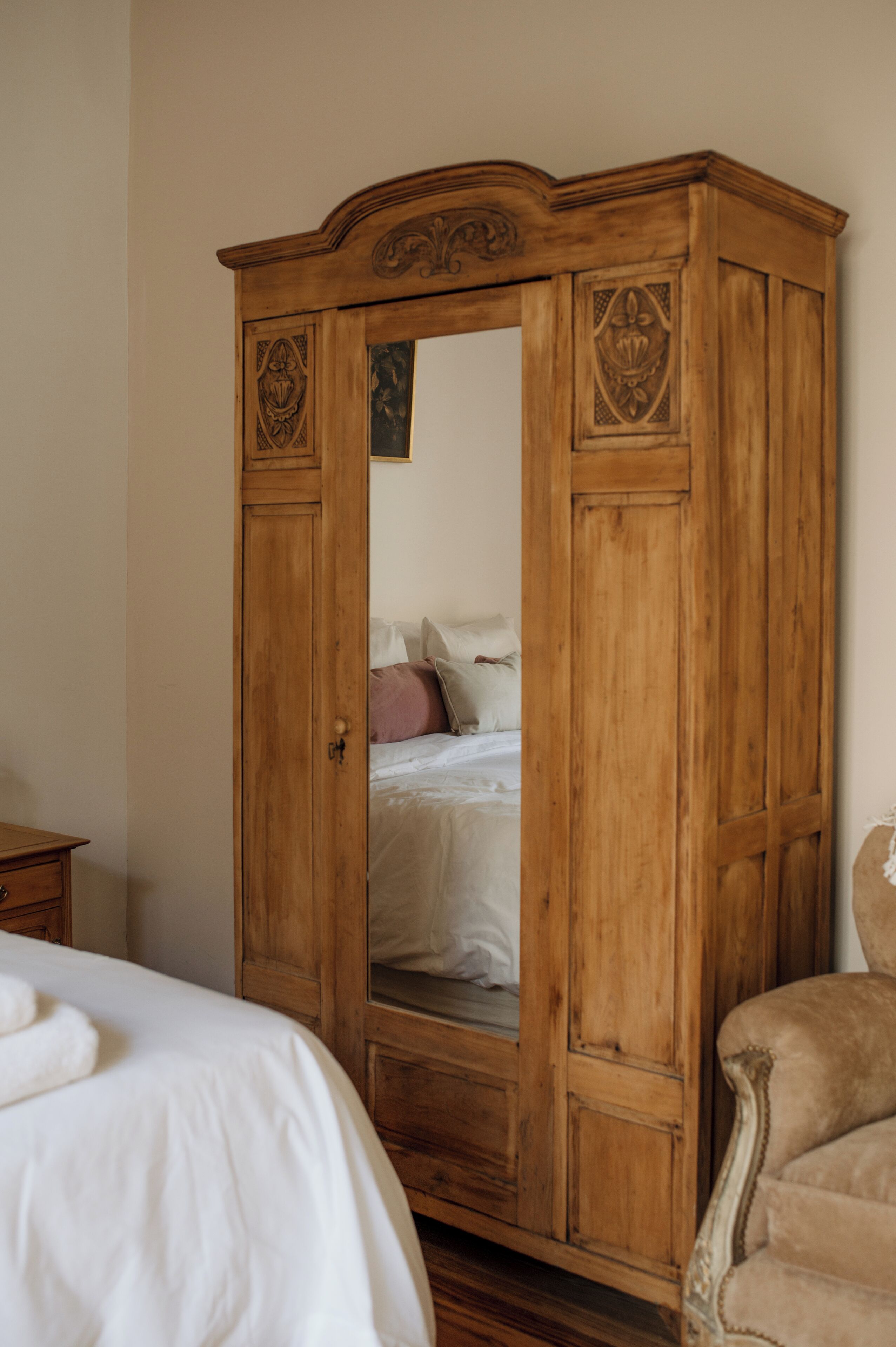 A wooden wardrobe with a mirror, a bed with white bedding, and a chair.