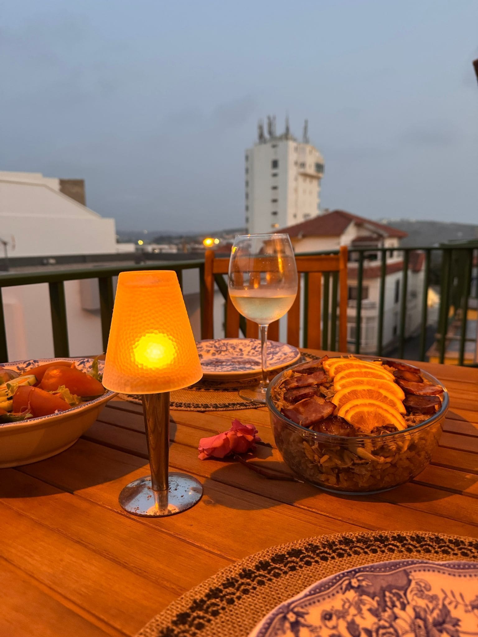 A table with a bowl of food, a wine glass, and a table lamp on a balcony overlooking a cityscape.