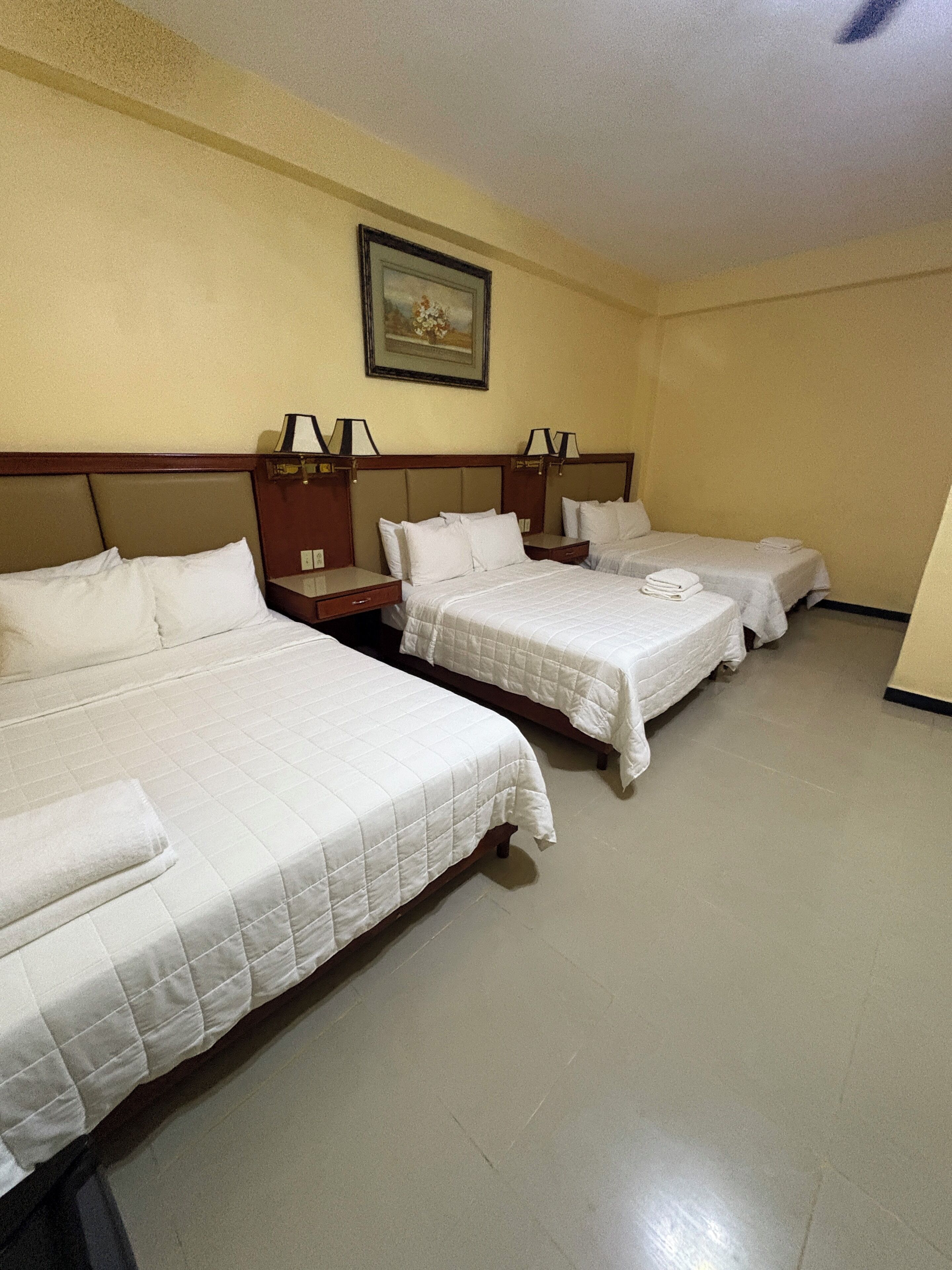 A hotel room with three beds, each with white bedding and a quilted cover. There is a painting on the wall above the beds and a small table with a lamp between them.