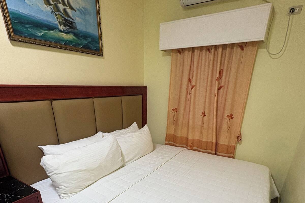 A neatly made bed with white linens, a headboard, a painting of a ship on the wall, and an air conditioning unit above the bed.