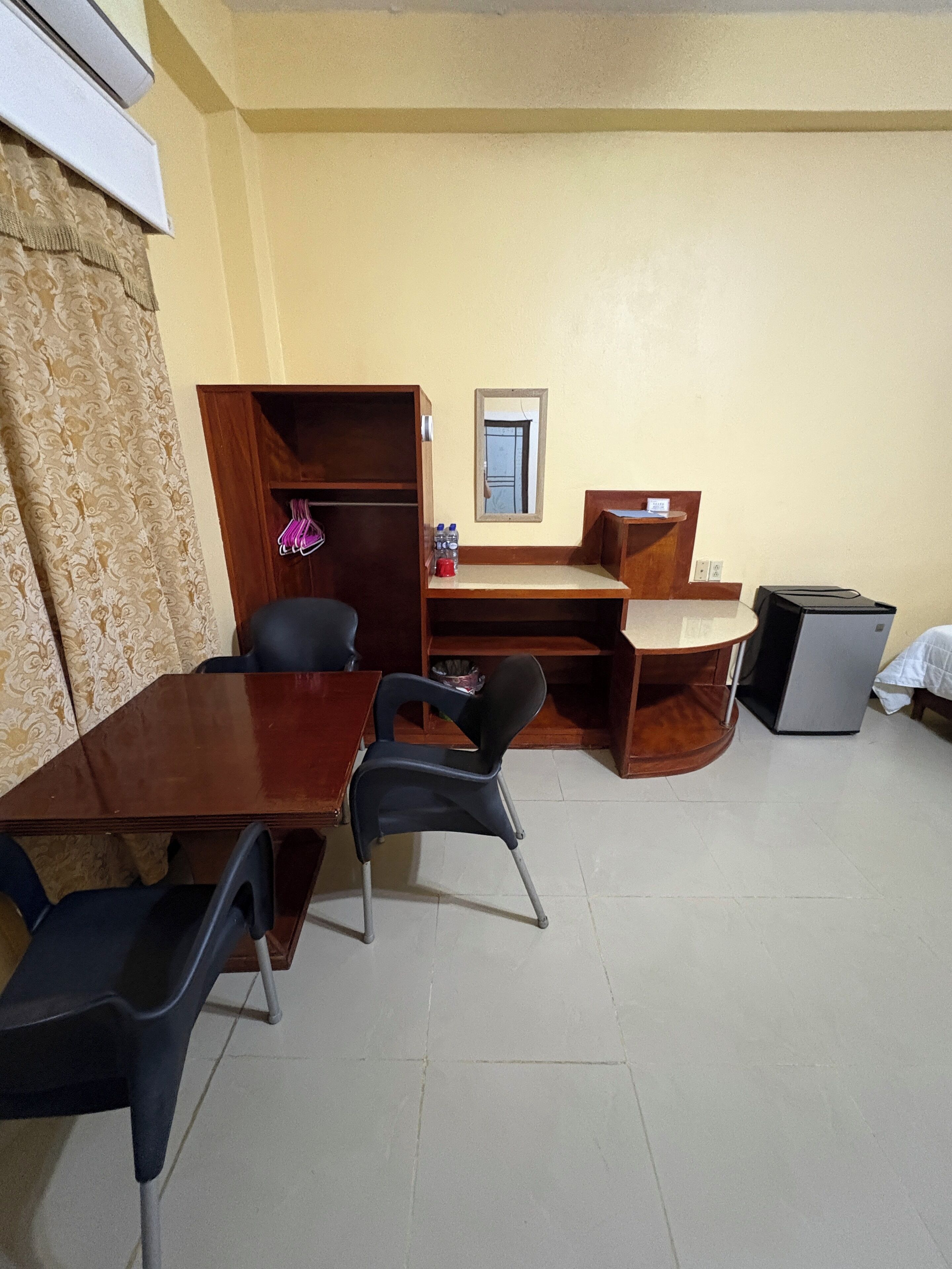Une pièce comprenant un bureau en bois, deux chaises, une petite armoire en bois, un miroir et une poubelle.