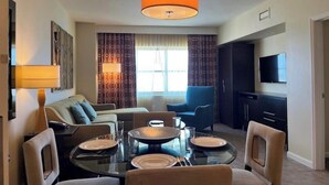 Een moderne woonkamer met een glazen eettafel voor vier personen, een bank, een blauwe fauteuil en een televisie aan de muur.