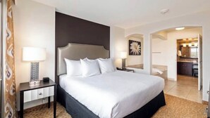Hotelkamer met een groot bed, nachtkastje met lamp, donker hoofdeinde, en zicht op de keuken en badkamer via een boog.