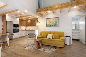 A modern kitchen with built-in appliances, a dining area with a wooden table and chairs, a yellow sofa, and a large painting on the wall.