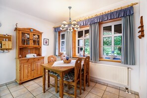 A dining area with a wooden table, chairs, a chandelier, a window with curtains, and a wooden cabinet with glass doors.
