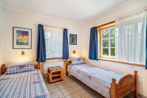 Quarto com duas camas de solteiro, uma mesinha, um radiador e duas janelas com cortinas.