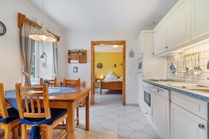 A compact kitchen with wooden cabinets, a dining area with a table and chairs, and a bedroom visible through an open door.
