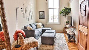 A living room with a gray sectional sofa, a patterned armchair, a gray ottoman, a large mirror, a floor lamp, a potted plant, and a wooden cabinet. - Lookout Loft over the Monroe Square (Monroe)