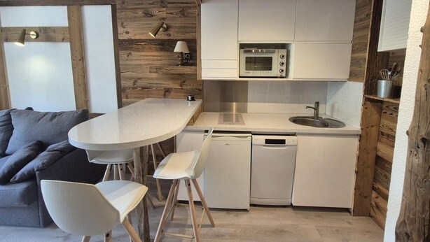 Een compacte keuken met witte kastjes, een inbouwoven en een vaatwasser. In de aangrenzende woonkamer staat een rond tafel met twee stoelen en een grijze bank.