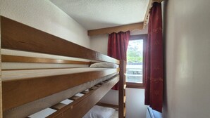 Een stapelbed met houten frame en wit beddengoed, een raam met rode gordijnen en een radiator aan de muur.
