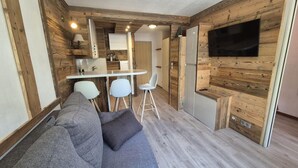 Een ruimtelijk ingerichte leefruimte met een bank, een eettafel met stoelen, een televisie aan een houten wand en een kitchenette.
