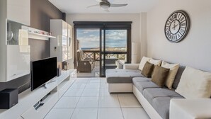 Ein modernes Wohnzimmer mit Flachbildfernseher, Deckenventilator, großem Fenster und Blick ins Freie.