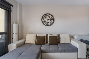 Un salon moderne avec un canapé d’angle, une horloge murale et une fenêtre équipée de stores.