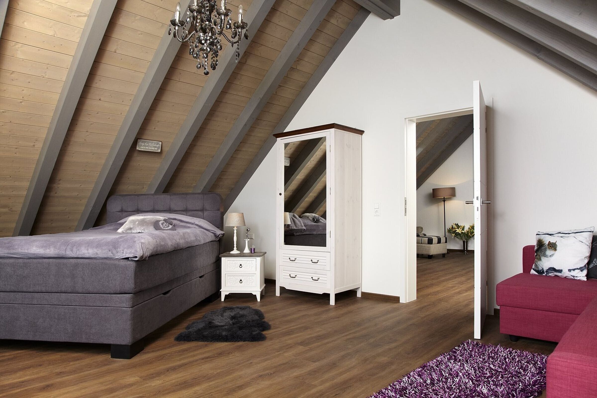 A bedroom with a sloped ceiling, a bed, a wardrobe, a nightstand, a lamp, a red sofa, and a cat.