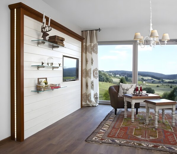 A modern living room with a large window, a dining area, and a TV mounted on the wall. - Apartment 'Kuckucks' with Spa Area and Mountain View (Ibach)